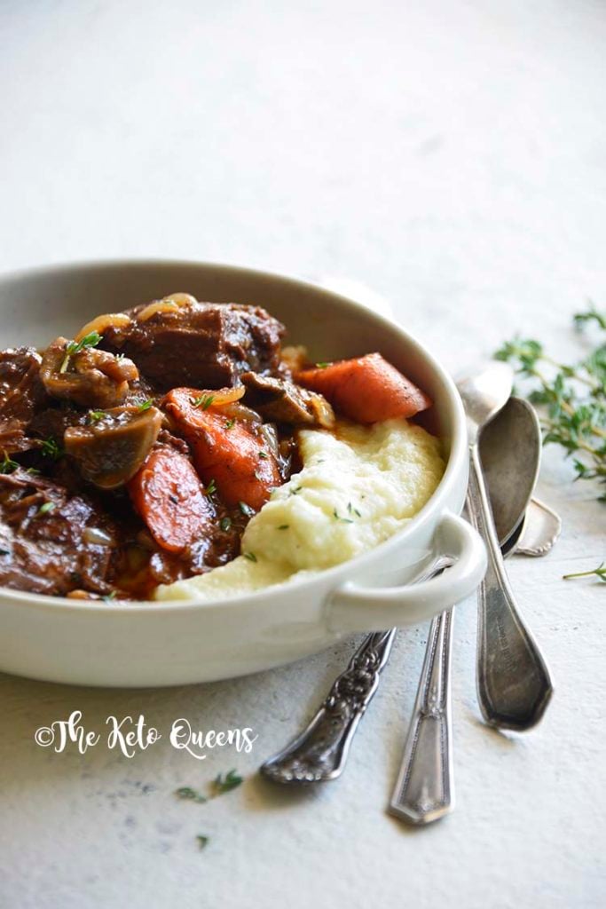 vertical 3/4 view closeup image of low carb instant pot beef stew in a white bowl with 3 spoons and fresh herbs on a white background